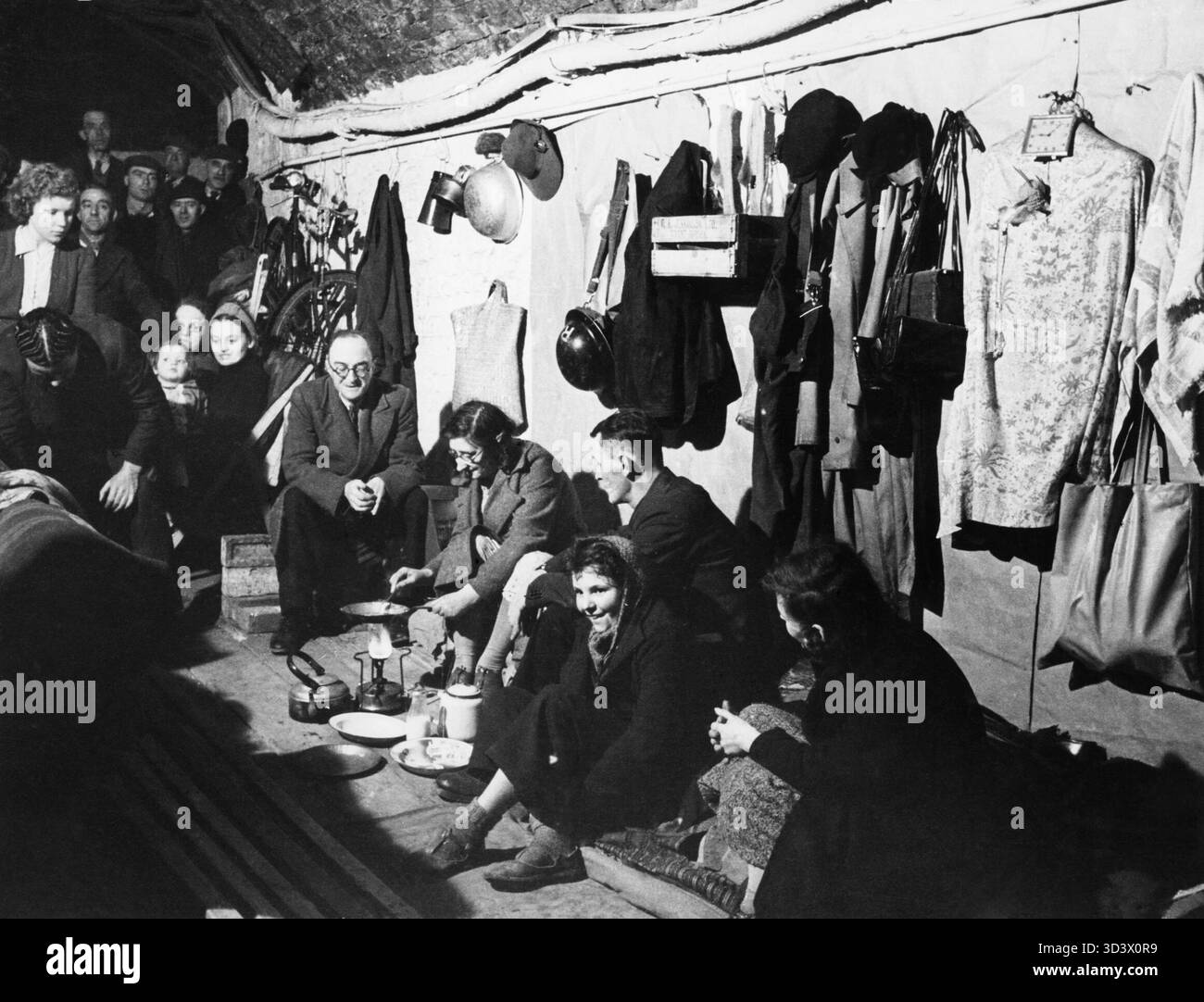 A woman cooks a meal over a Primus stove in a public air raid shelter under railway arches in South East London, 1940. A woman cooks a meal in a frying pan over a Primus stove whilst other shelterers look on in this shelter under the railway arches, somewhere in South East London. Many items of clothing and other personal possessions, including hats and gas mask boxes, have been hung on nails along the whitewashed walls of the shelter. This photograph was probably taken in November 1940. Stock Photo