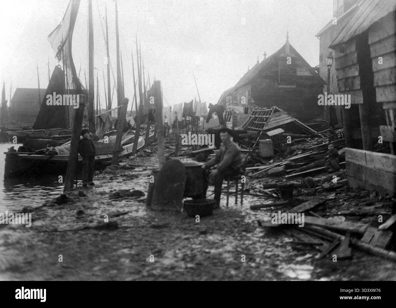 This image shows the aftermath of the 1916 flooding disaster in Spakenburg, Netherlands. A man is seen holding a chair and a feed kettle, his only possessions, with sailboats, destroyed houses, and clothes on a clothesline in the background. Stock Photo