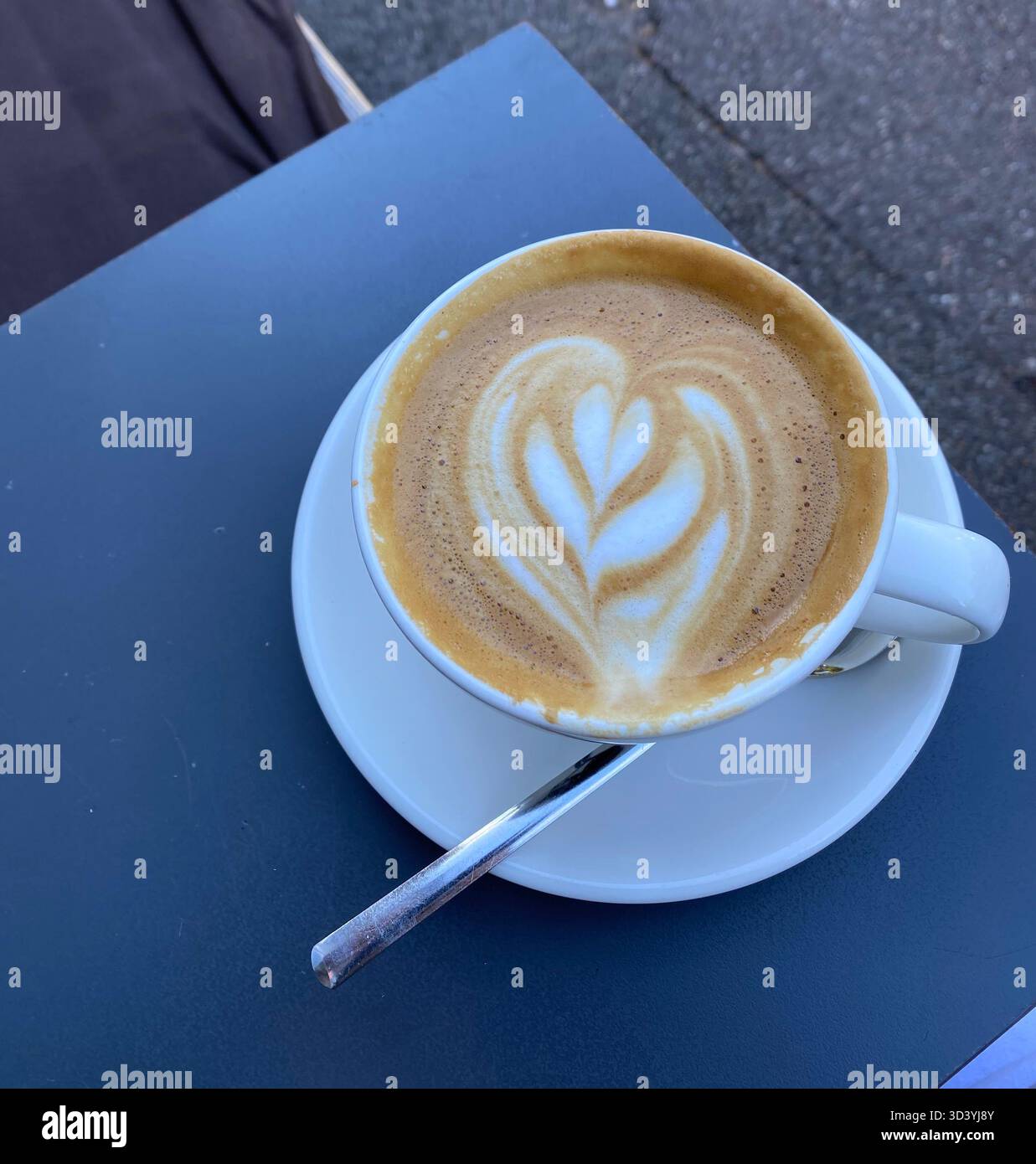 Cup of cappuccino with heart latte art in a white cup and saucer with spoon on an outdoor café table, top view. - Smartphone Captured Stock Image