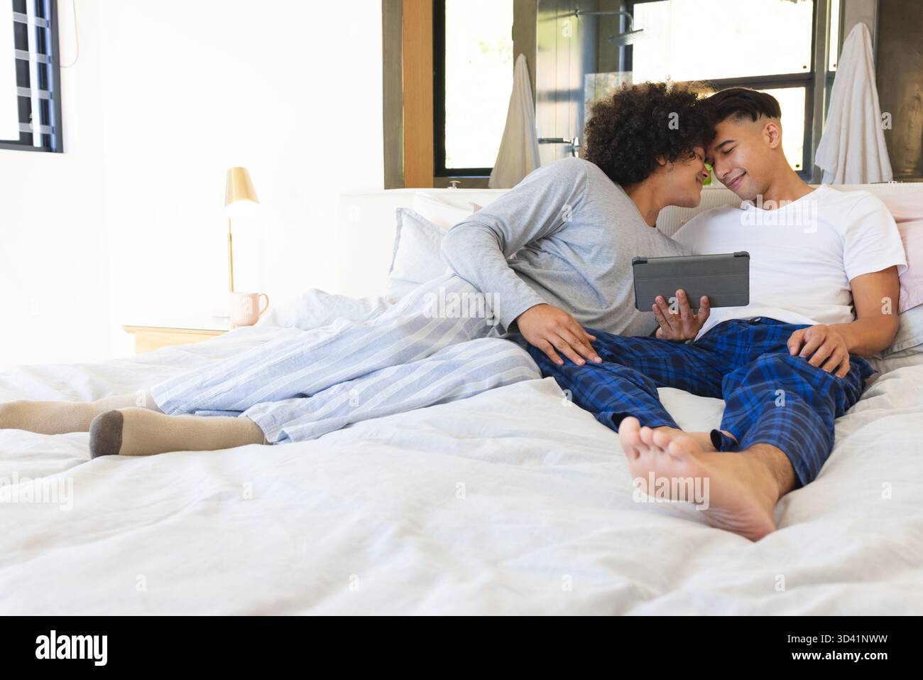Relaxing together two men sharing tablet on bed in bright modern bedroom, with pillows and blanket Stock Photo