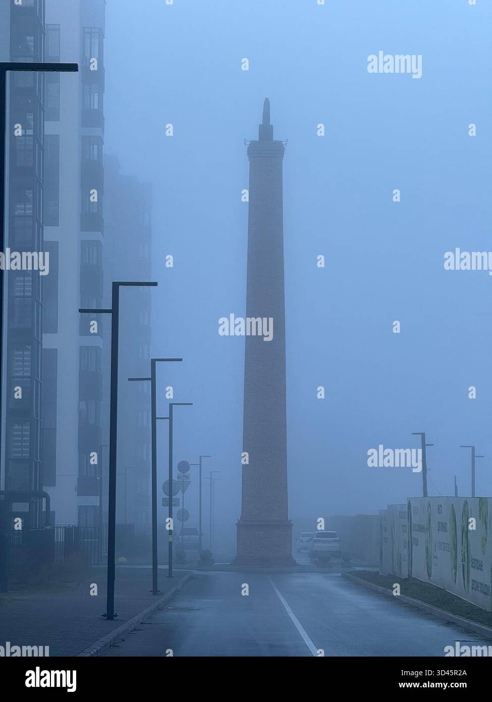 A foggy street with a tall stone obelisk in the center, wet road leading toward it, and streetlights and houses fading into the dense white mist. - Smartphone Captured Stock Image