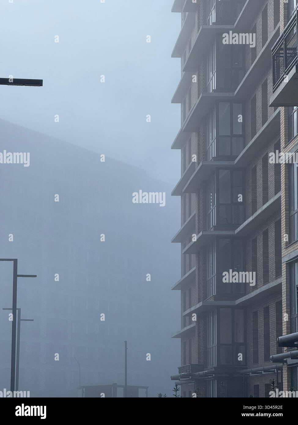 A foggy street with a brown brick building on the right side and dense white mist covering the left side, creating a calm and mysterious atmosphere. - Smartphone Captured Stock Image