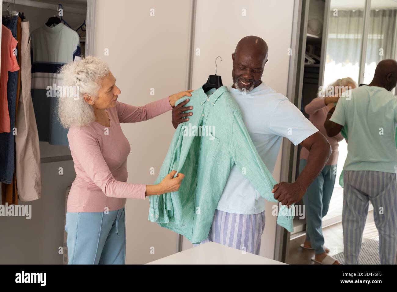 Holding Diverse couple trying on light green shirt in bedroom area, with wardrobe and mirror Stock Photo
