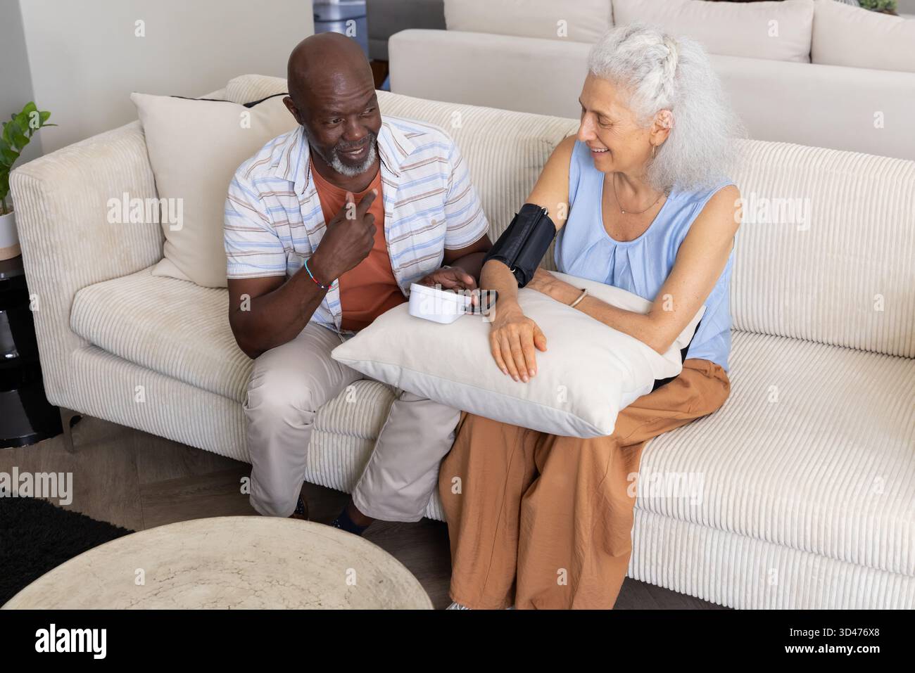 Monitoring diverse senior couple using blood pressure monitor in living room, with pillow Stock Photo