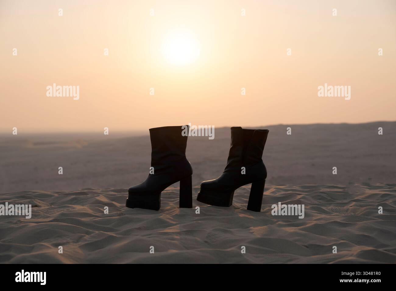 Doha, Qatar - October 30, 2025: A pair of stylish black high-heeled boots standing on soft desert sand during a serene sunset. Stock Photo