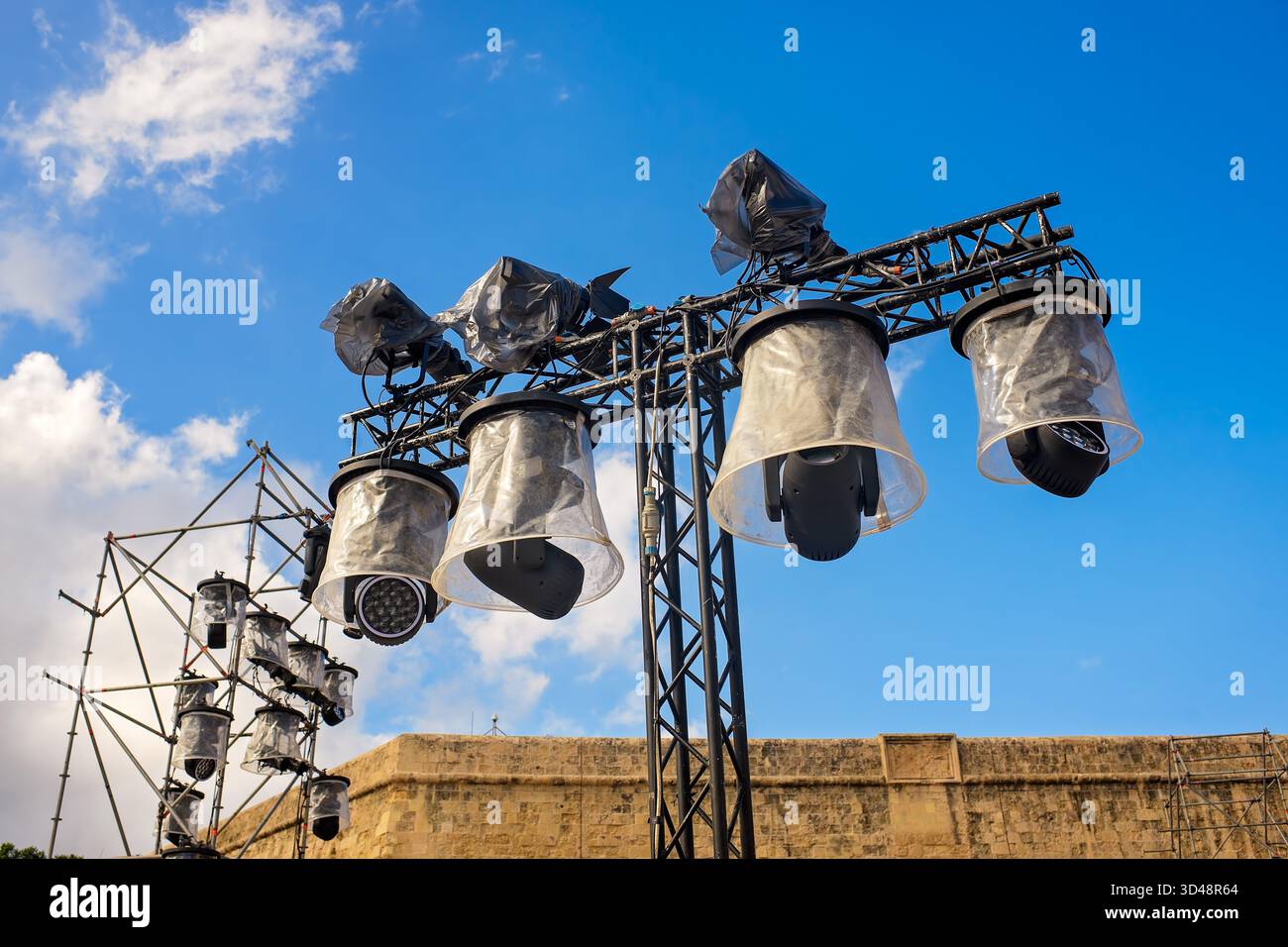 Professional stage lights installed on trusses before an outdoor event, covered with plastic and tape for weather protection under a blue sky. Stock Photo