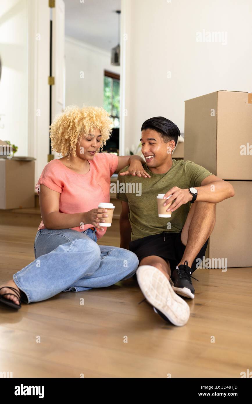 Young diverse couple sitting on floor with coffee cups, enjoying new home together. Moving, relocation, new beginnings, relaxing, settling, residential || Model released Stock Photo