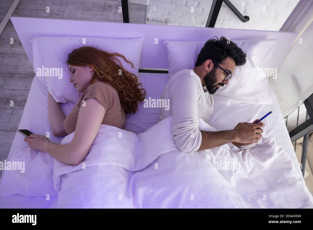 Young couple in bed using phone lying backs to each other Stock Photo