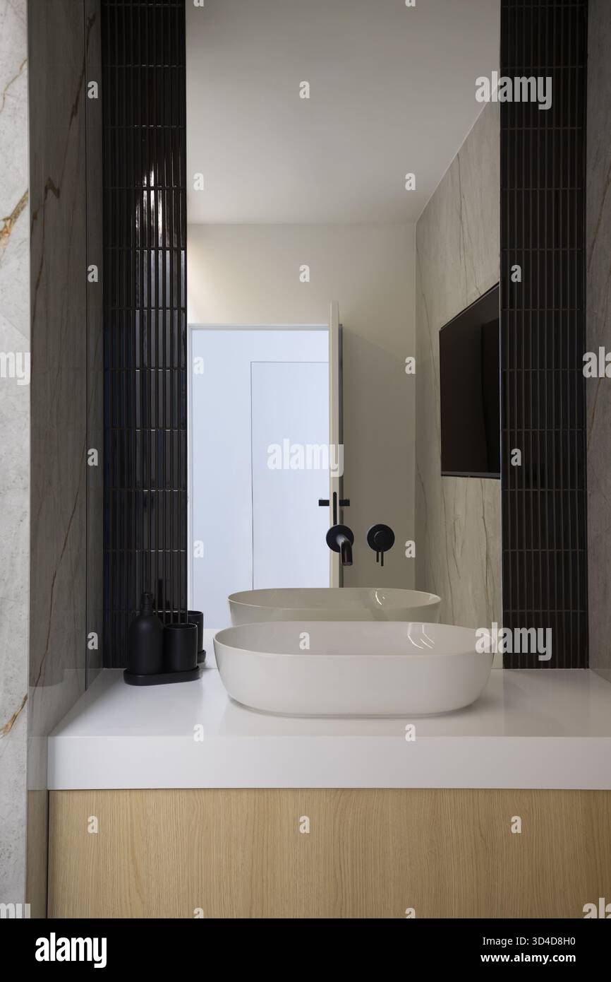 Interior of a modern and luxurious bathroom. It features a white ceramic vessel sink on a white countertop, placed on a wooden vanity. The walls are d Stock Photo