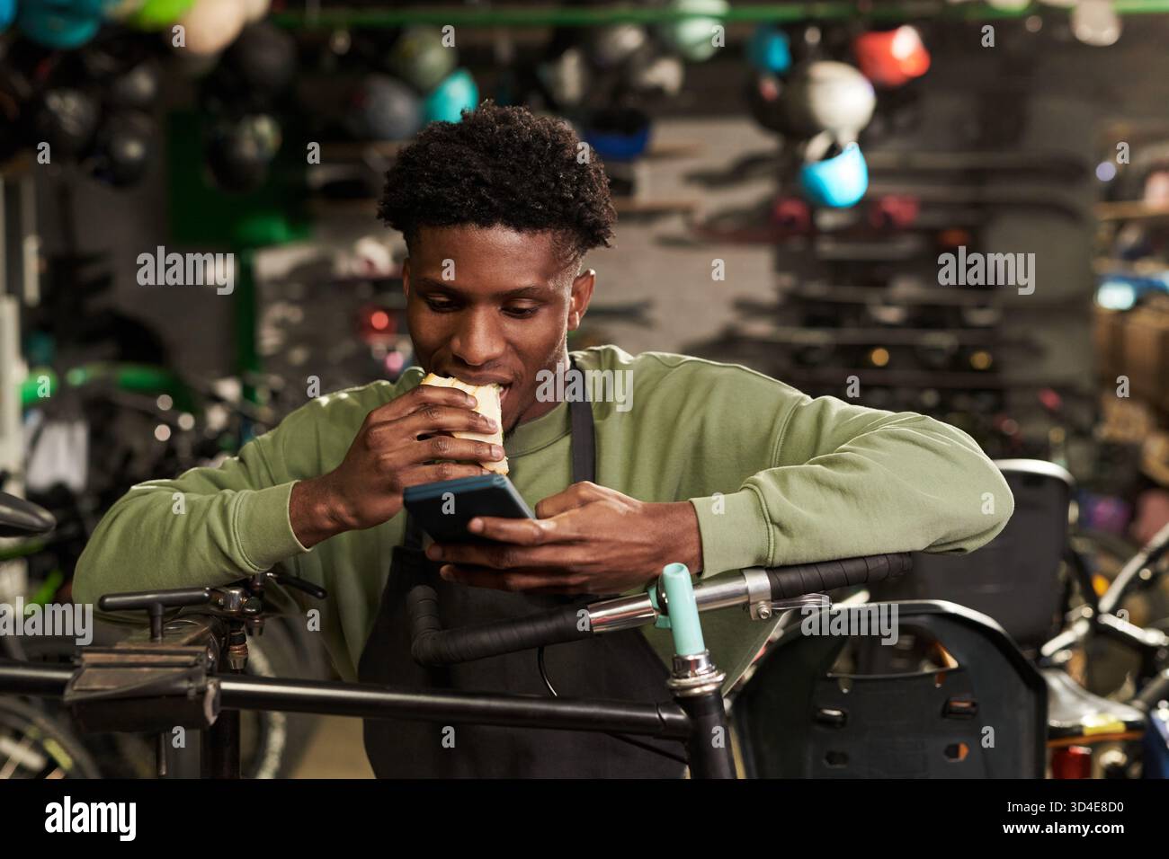 Young Black Man Eating Sandwich While Using Smartphone in Workshop Stock Photo