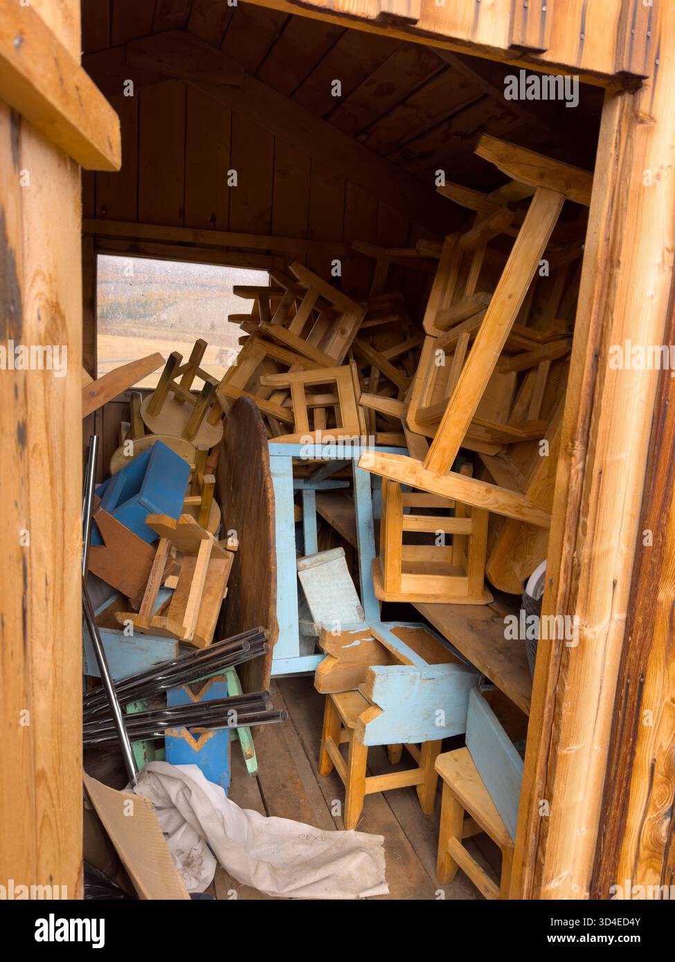 Chaotic scene inside wooden shed packed with chairs tables and planks stacked haphazardly. Weathered blue furniture contrasts warm wood tones creating rustic storage vibe. Stock Photo