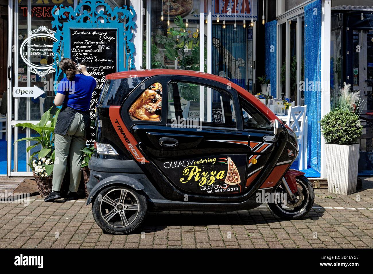 Pizza delivery microcar parked outside a restaurant, with a waitress writing the menu on the blackboard. Stock Photo