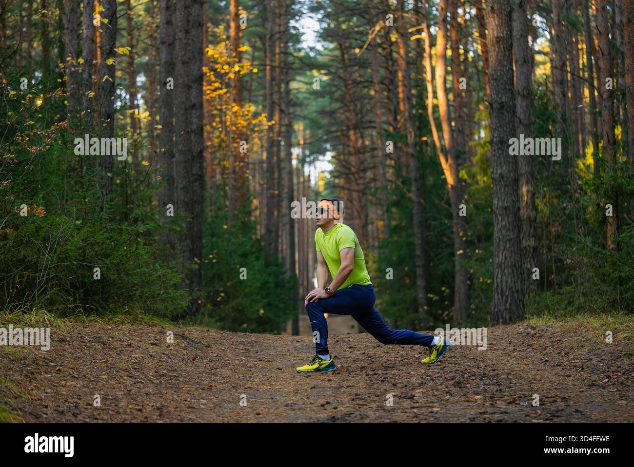 Male runner in sportswear performing a lunge stretch on a forest path, focusing on warming up muscles and maintaining a healthy lifestyle while exercising in nature during autumn Stock Photo