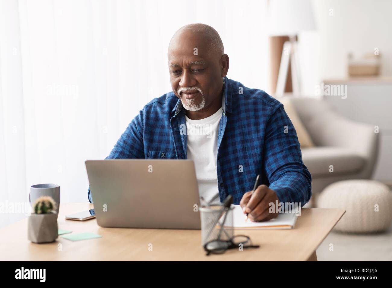 Senior Black Man Working at Home Using a Laptop and Taking Notes While Learning Online Stock Photo