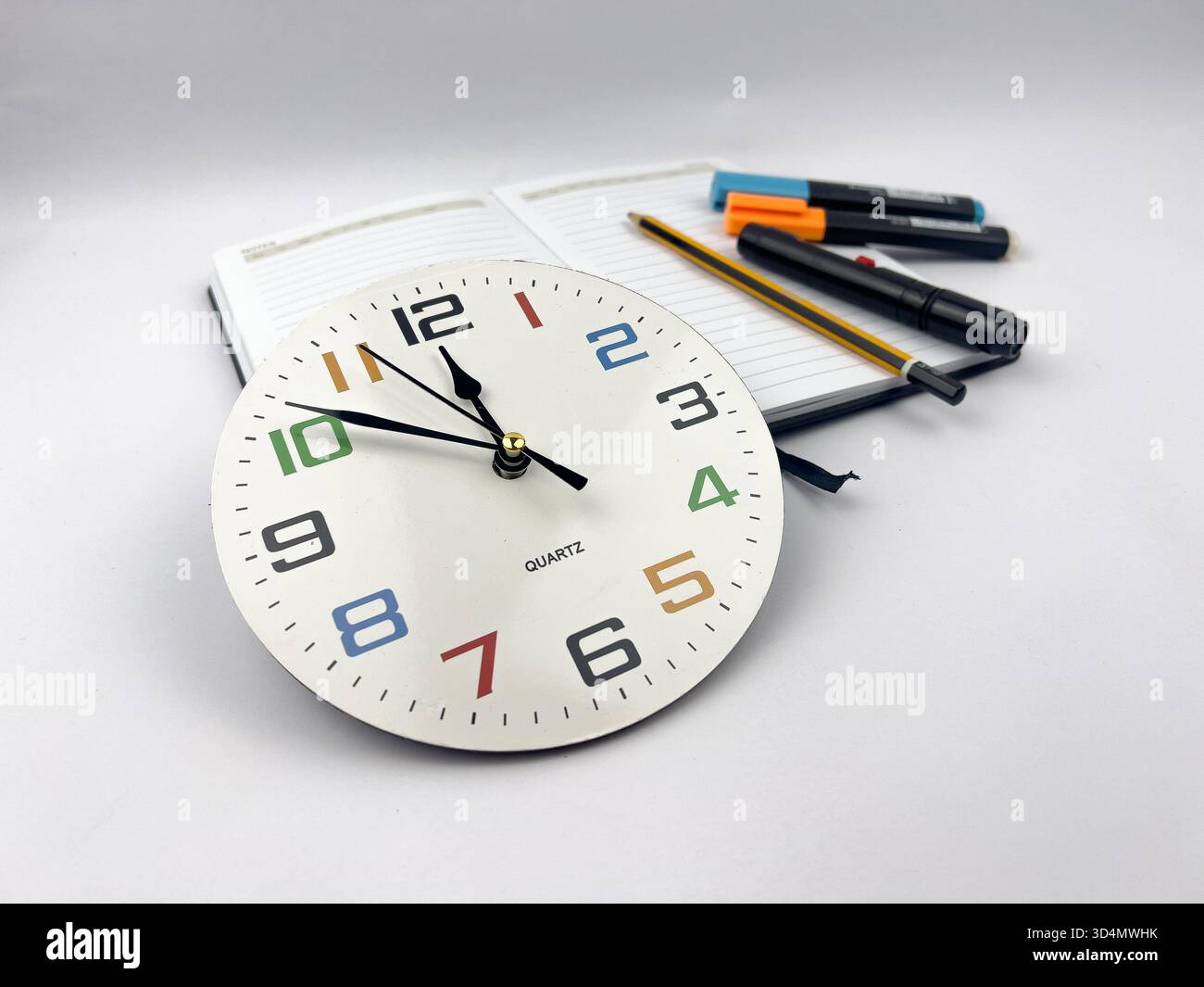 A still life composition centered around a clock face, a notebook, and various writing/marking tools. Stock Photo