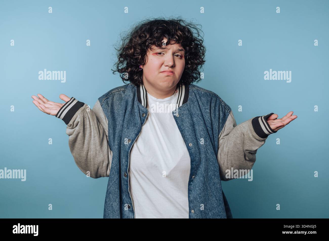 A person with curly hair is standing against a light blue background. They wear a denim jacket over a white shirt and have their hands raised in a ges Stock Photo