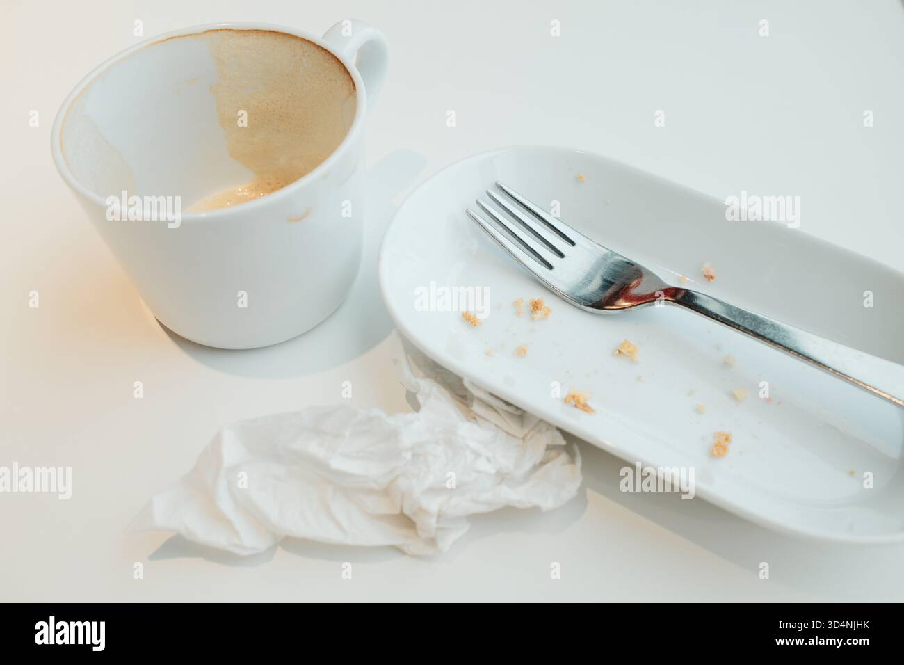 Empty cup with coffee foam, white plate with crumbs, fork, and crumpled napkin after a meal. Stock Photo