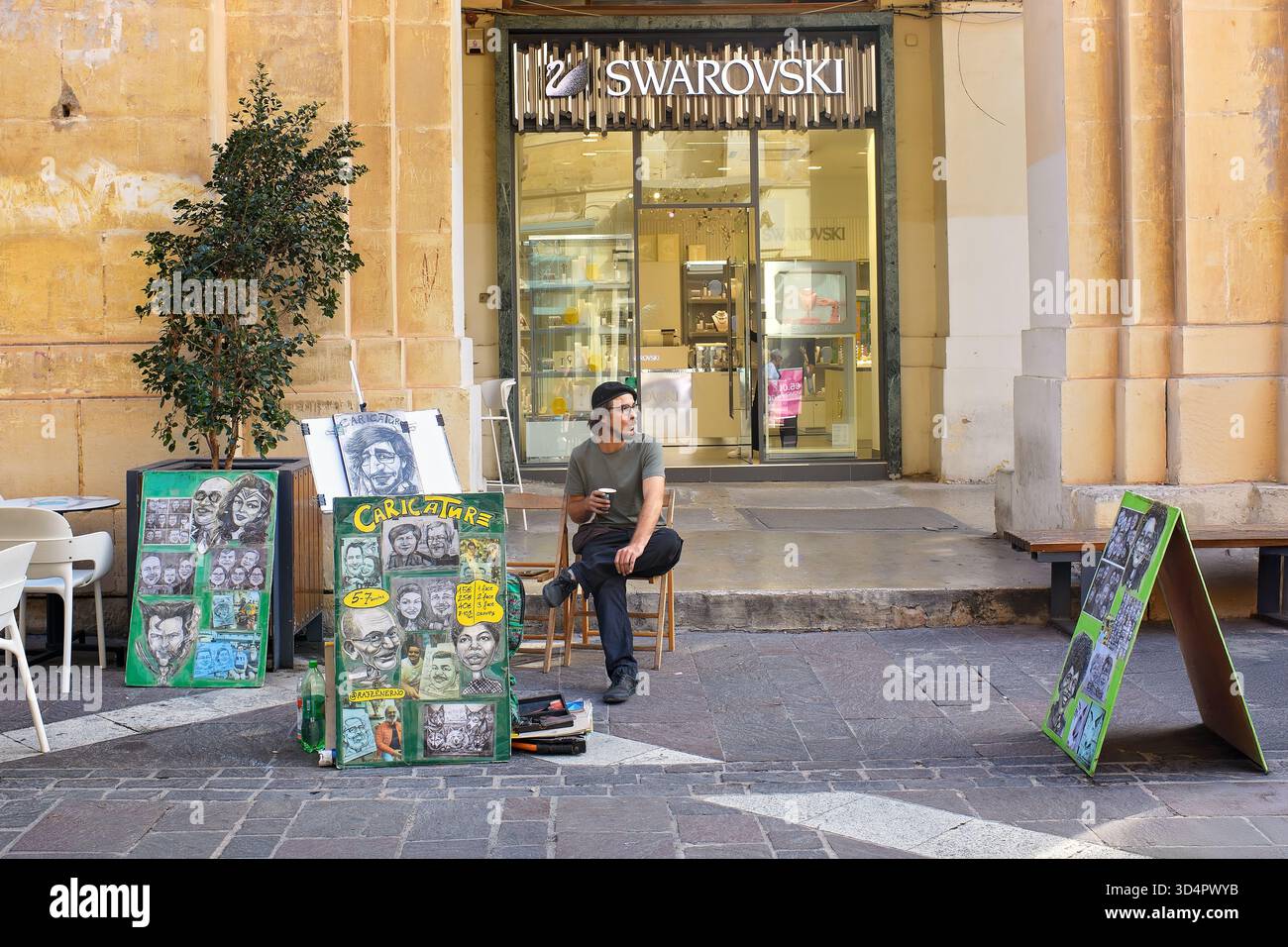 Valletta, Malta - 11.05.2025: A street artist in a black beret holding a cup of coffee sits with caricatures displayed outside a Swarovski store Stock Photo