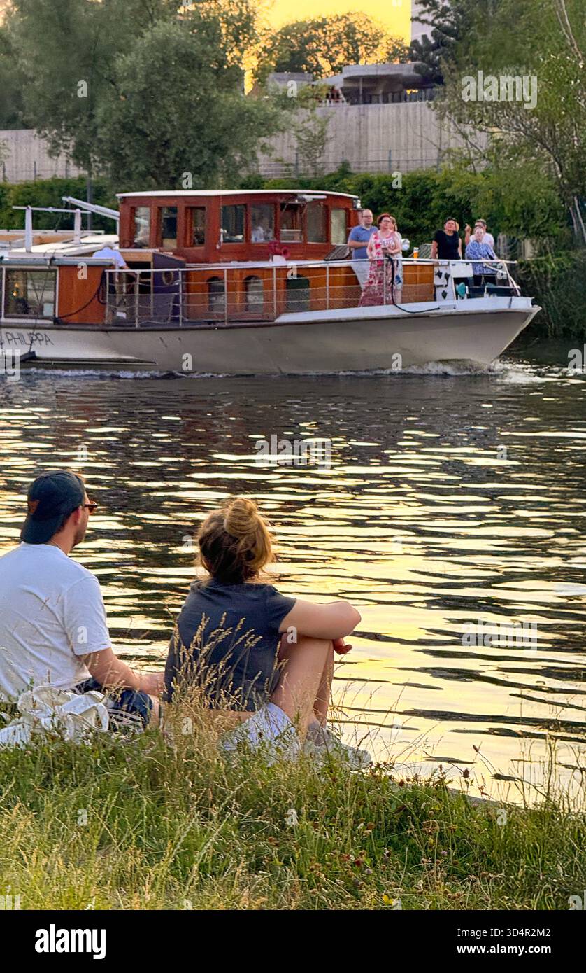 Berlin, Deutschland – Paar sitzt am Landwehrkanal und beobachtet ein Boot im Abendlicht Stock Photo
