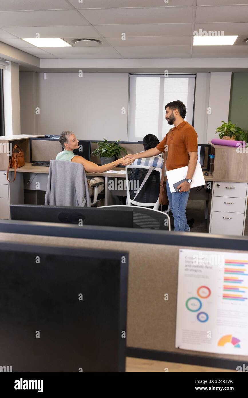 Shaking hands Diverse coworkers standing in open-plan cubicles, with computer monitors and yoga mat Stock Photo
