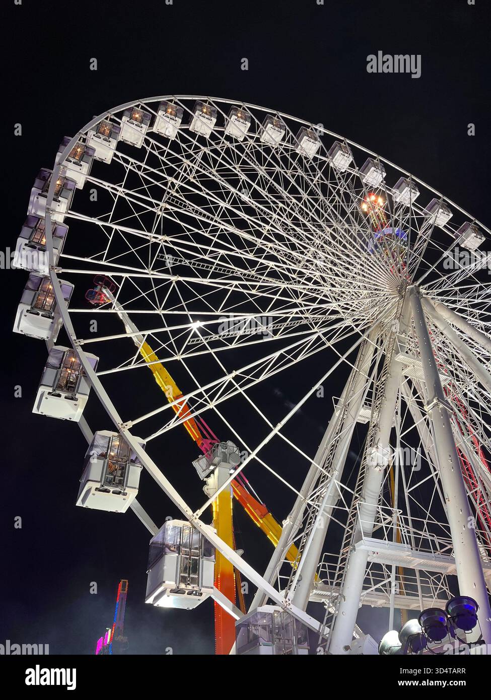 A brightly illuminated Ferris wheel captured from a low angle at night during a fair in Hull, East Yorkshire. The structure glows with colorful lights - Smartphone Captured Stock Image