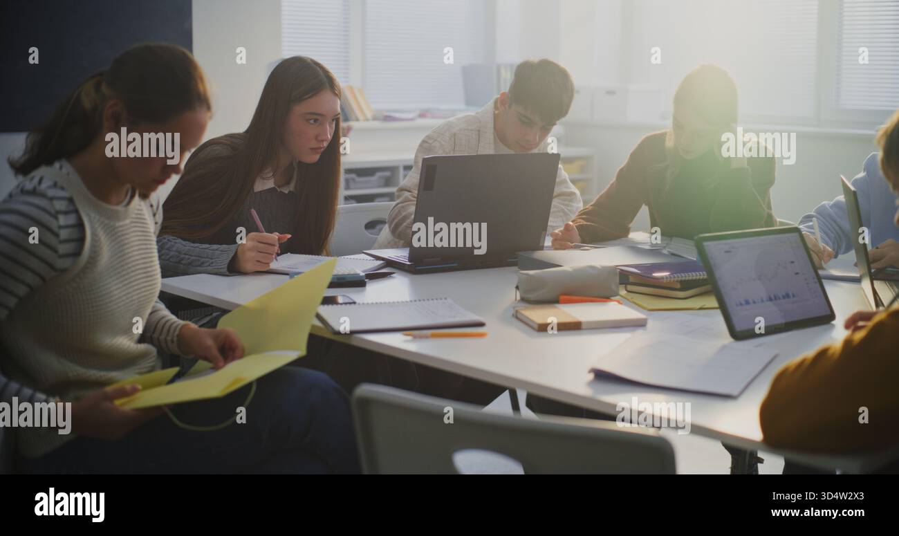 Group of Students Gathered Around Shared Workspace, Actively Collaborating on Assignment Using Laptops, Tablets, and Paper Notes. Concept of Cooperative Problem Solving, and Modern Classroom Teamwork. Stock Photo