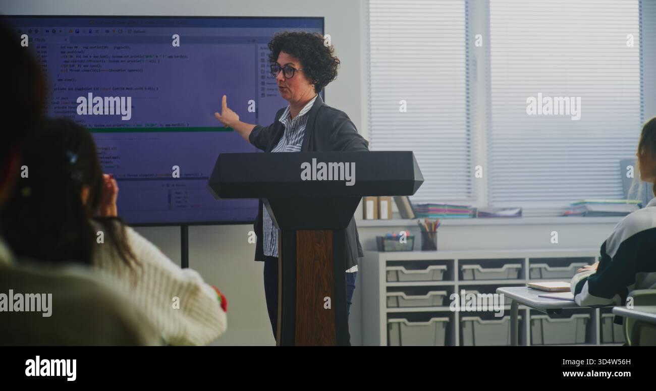 Female Coding Teacher Stands at Podium, Explaining Programming Syntax on Large Digital Screen. Concept Modern Computer Science Education, STEM Learning, Technology Literacy, and Teaching Innovation. Stock Photo