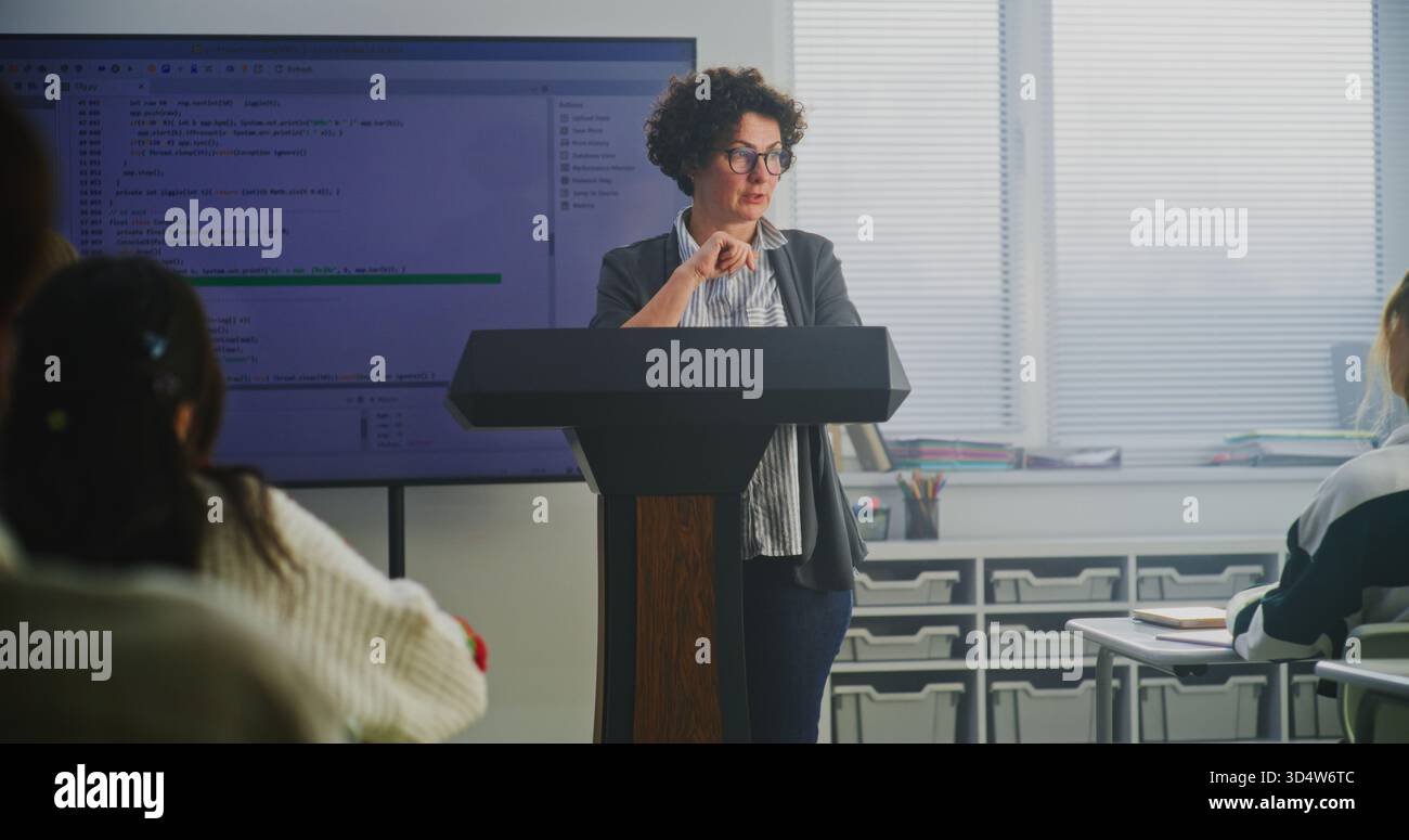 Female Coding Teacher Stands at Podium, Explaining Programming Syntax on Large Digital Screen. Concept Modern Computer Science Education, STEM Learning, Technology Literacy, and Teaching Innovation. Stock Photo