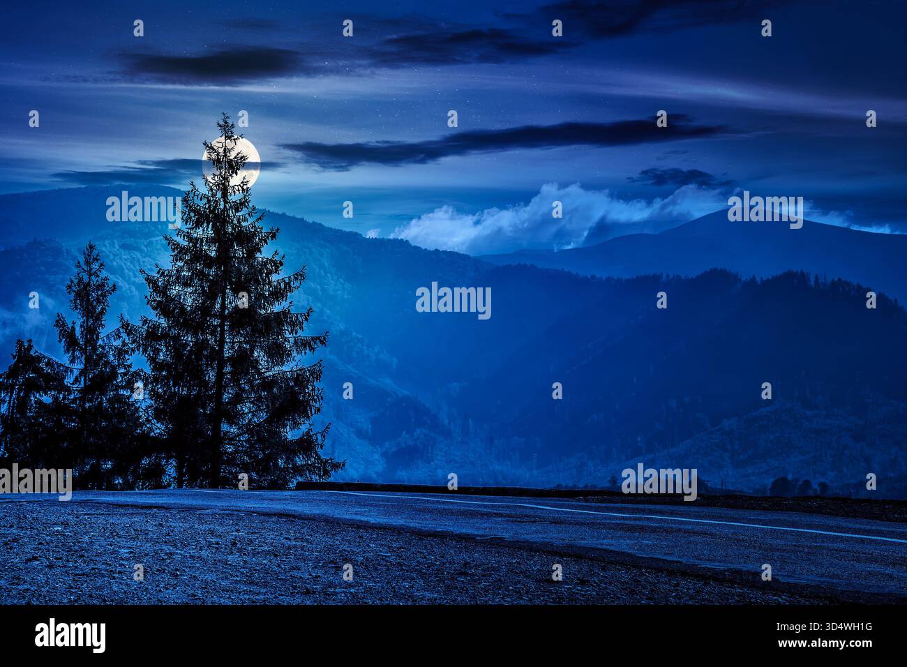 old asphalt road going through the hillside and passes green spruce forest at night. beautiful landscape with distant mountains in full moon light. ba Stock Photo