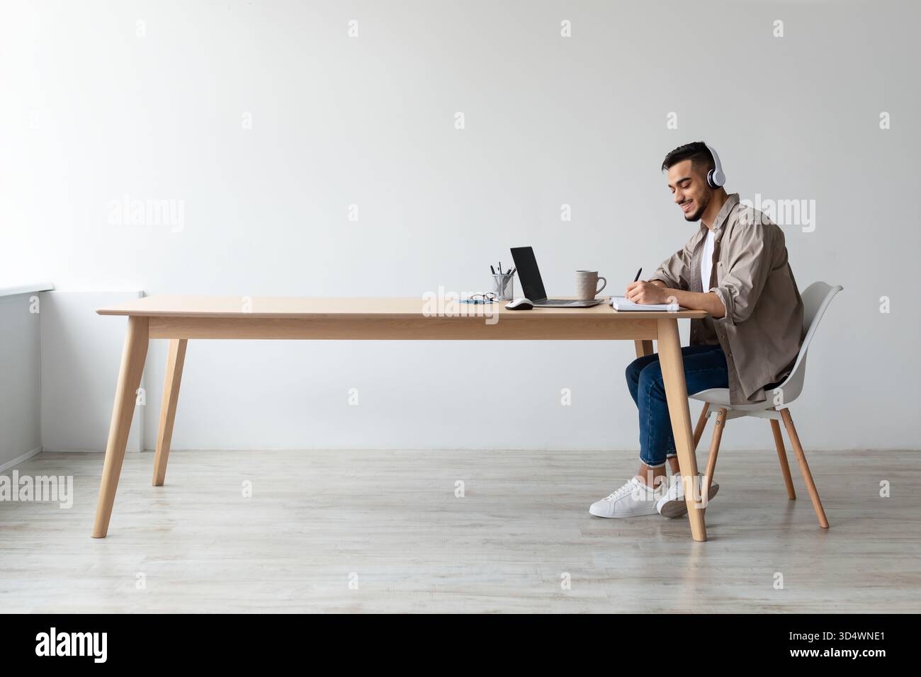 Smiling Arab Man Studies Online While Writing Notes and Using Laptop at Desk Stock Photo