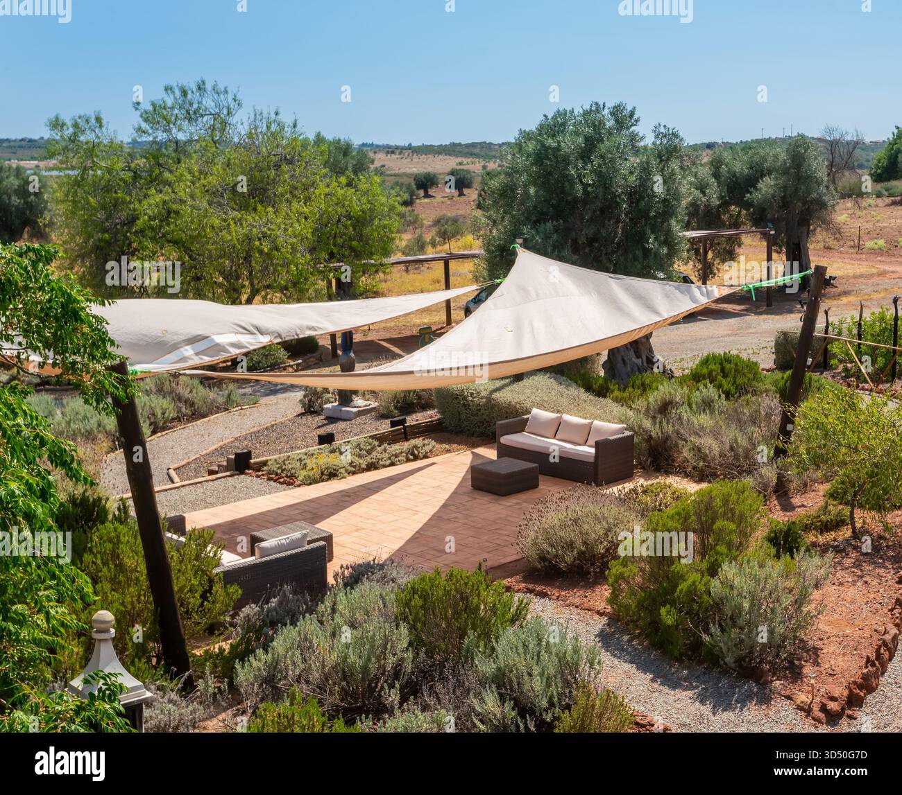 A peaceful outdoor patio features comfortable seating under shade sails, surrounded by greenery and open fields on a sunny day Stock Photo