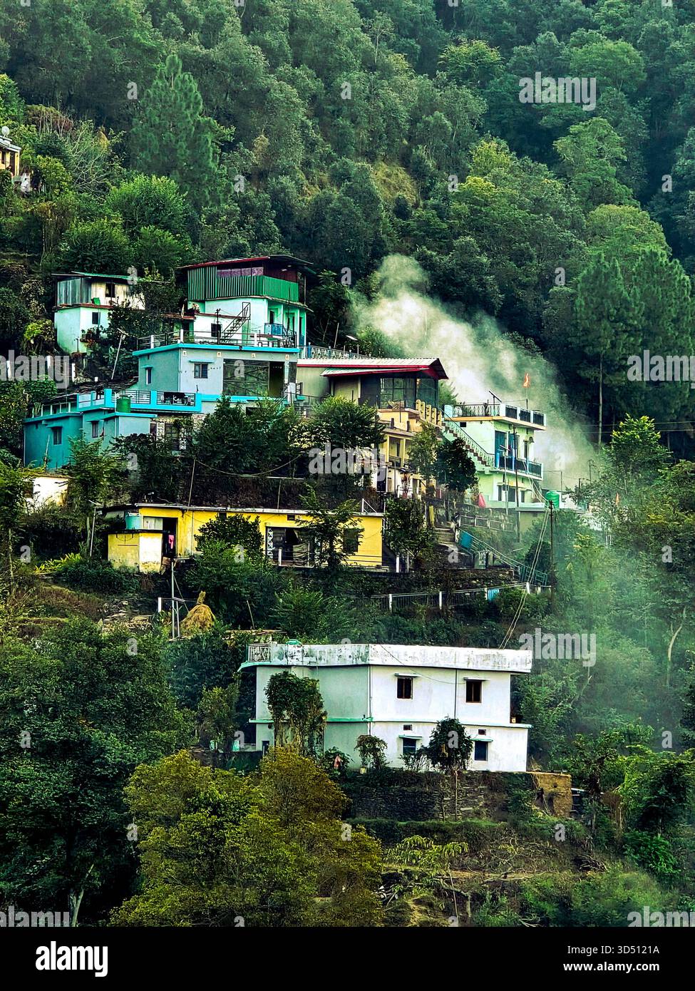 This image beautifully captures a peaceful hillside village nestled amidst dense green forests. The vibrant houses, painted in shades of turquoise, yellow, and white, stand gracefully on terraced slopes, creating a picturesque scene typical of the Himalayan or hilly regions of northern India. Wisps of smoke rise gently from one of the homes, suggesting early morning or evening cooking fires — a simple yet heartwarming symbol of daily rural life. The surrounding forest, filled with tall pine and dense foliage, wraps the village in a blanket of tranquility, making it feel almost hidden from the  - Smartphone Captured Stock Image