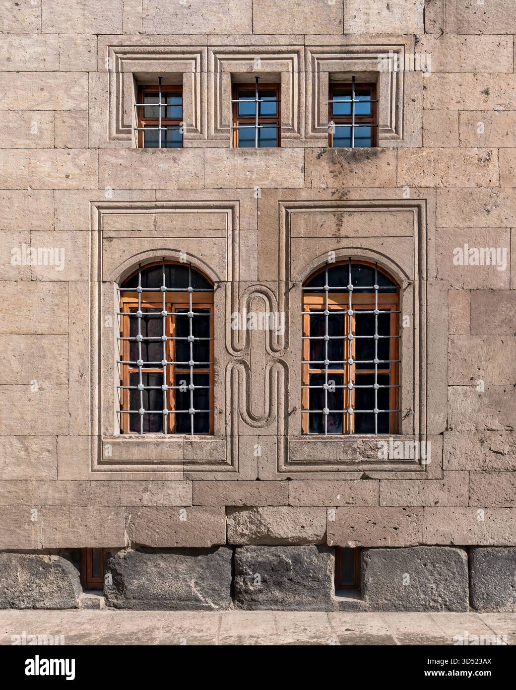 Stone facade with barred rectangular and arched windows, featuring carved frames and stylized geometric patterns. Stock Photo