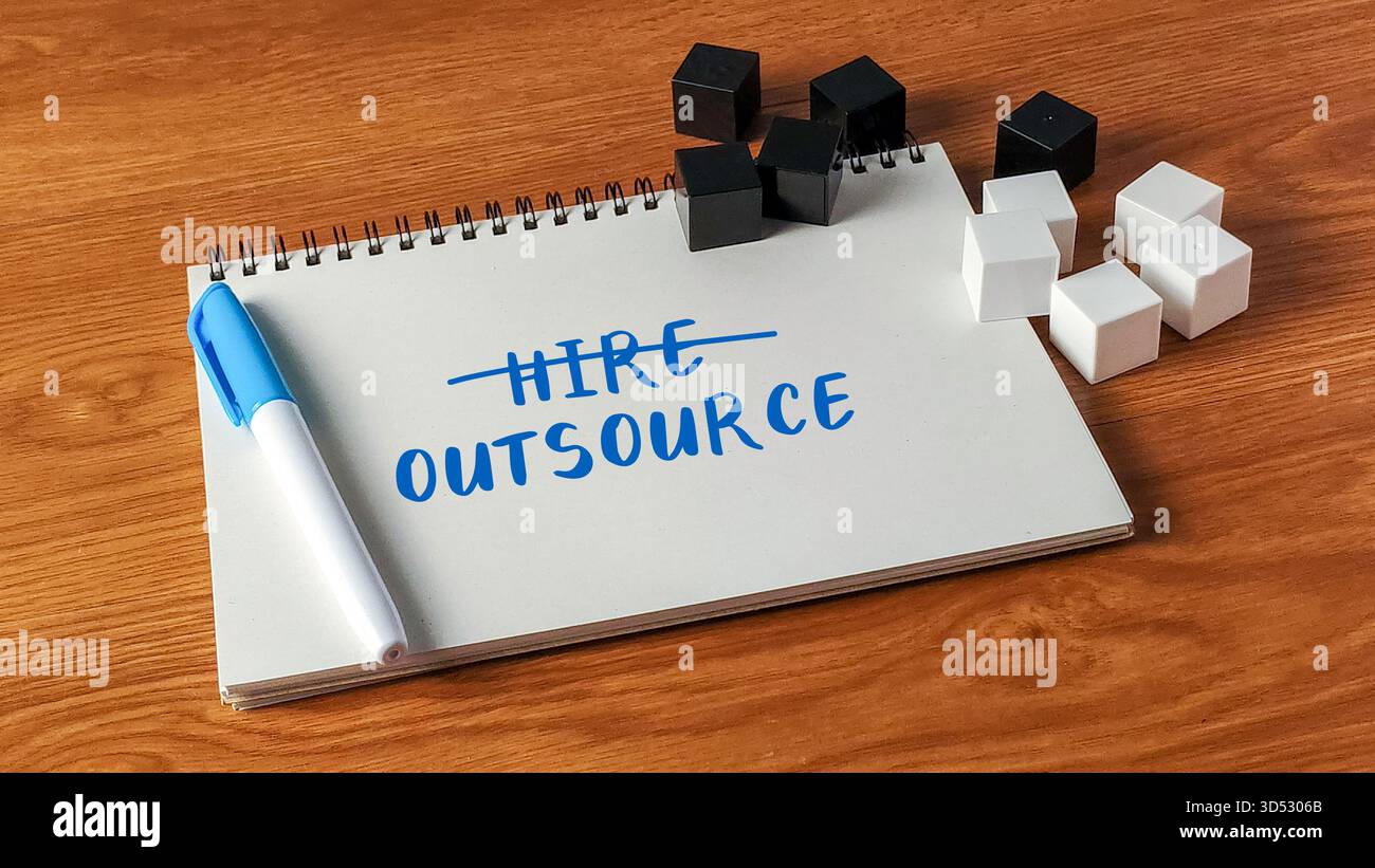 The word Hire crossed out and Outsource written on a notebook with blocks on a wooden desk symbolizing business strategy. Stock Photo