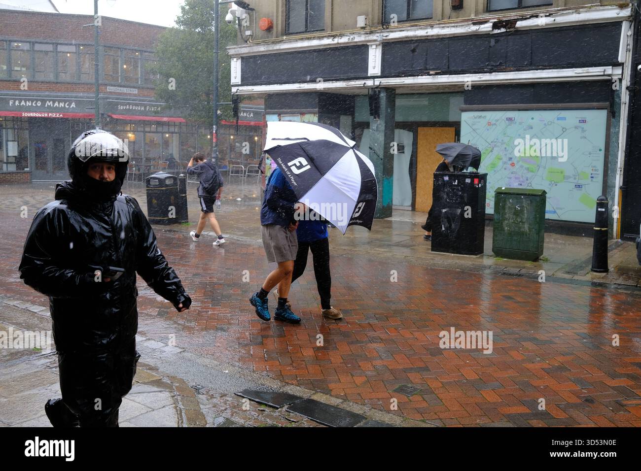 People shelter under a large black and white umbrella, while another wears a motorcycle crash helmet against the rains in Canterbury Kent 13/11/2025 Stock Photo