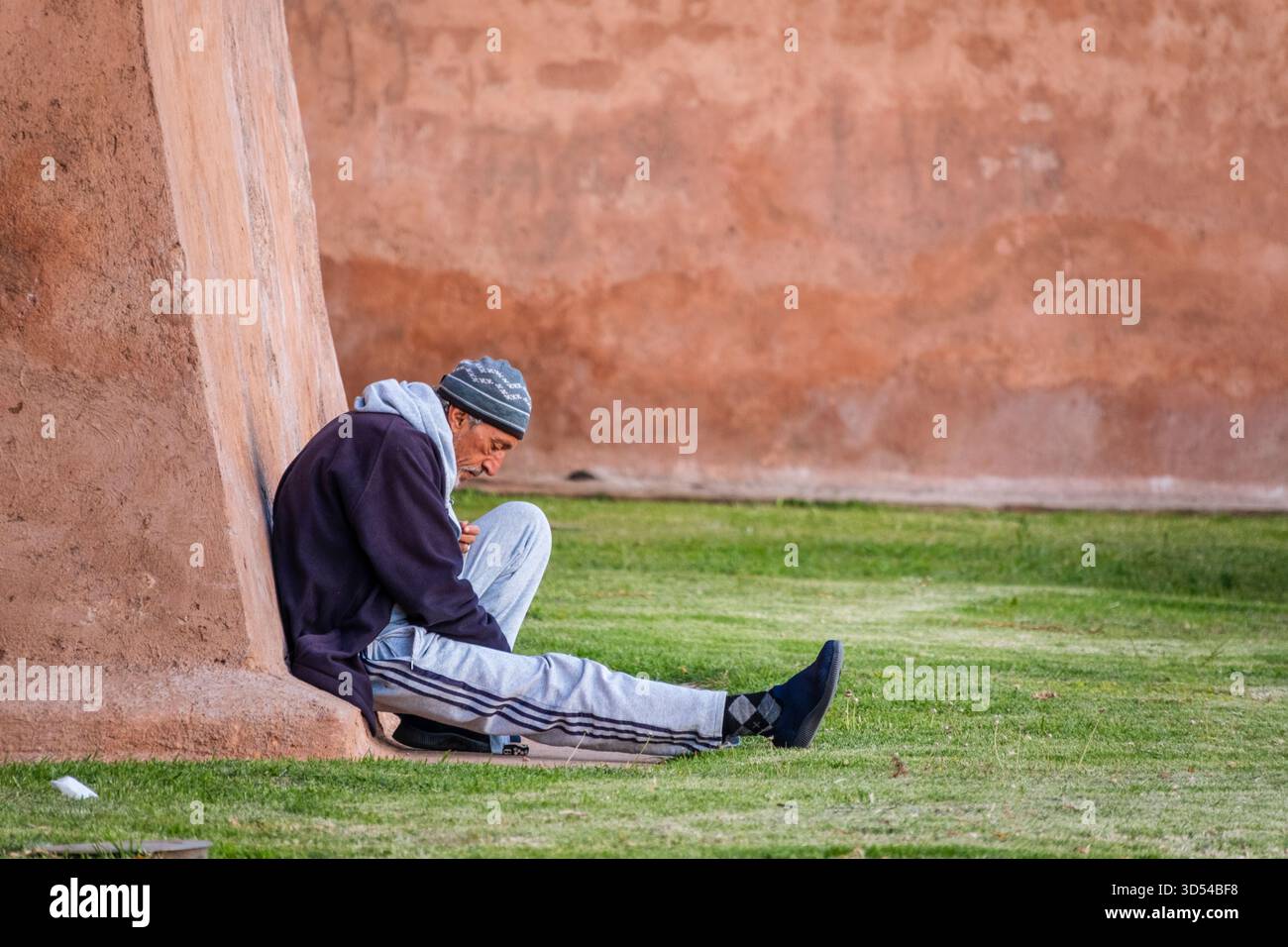Marrakesh, Morocco – April 21, 2025: an elderly man sits quietly against an old earthen wall in the historic medina. Dressed warmly in a beanie and ja Stock Photo