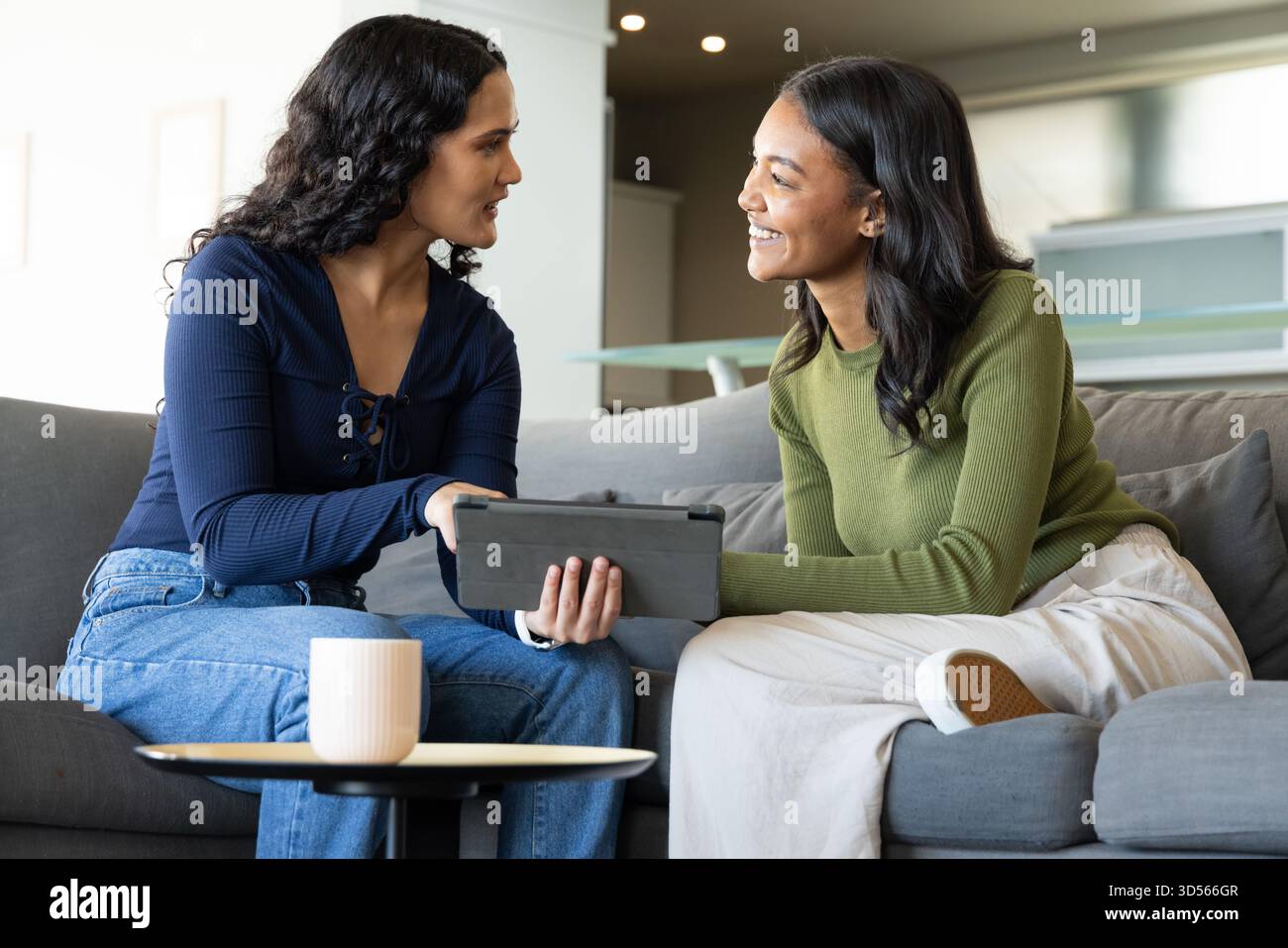 Sharing Diverse female friends browsing tablet on gray sofa in lounge, with coffee mug on table Stock Photo