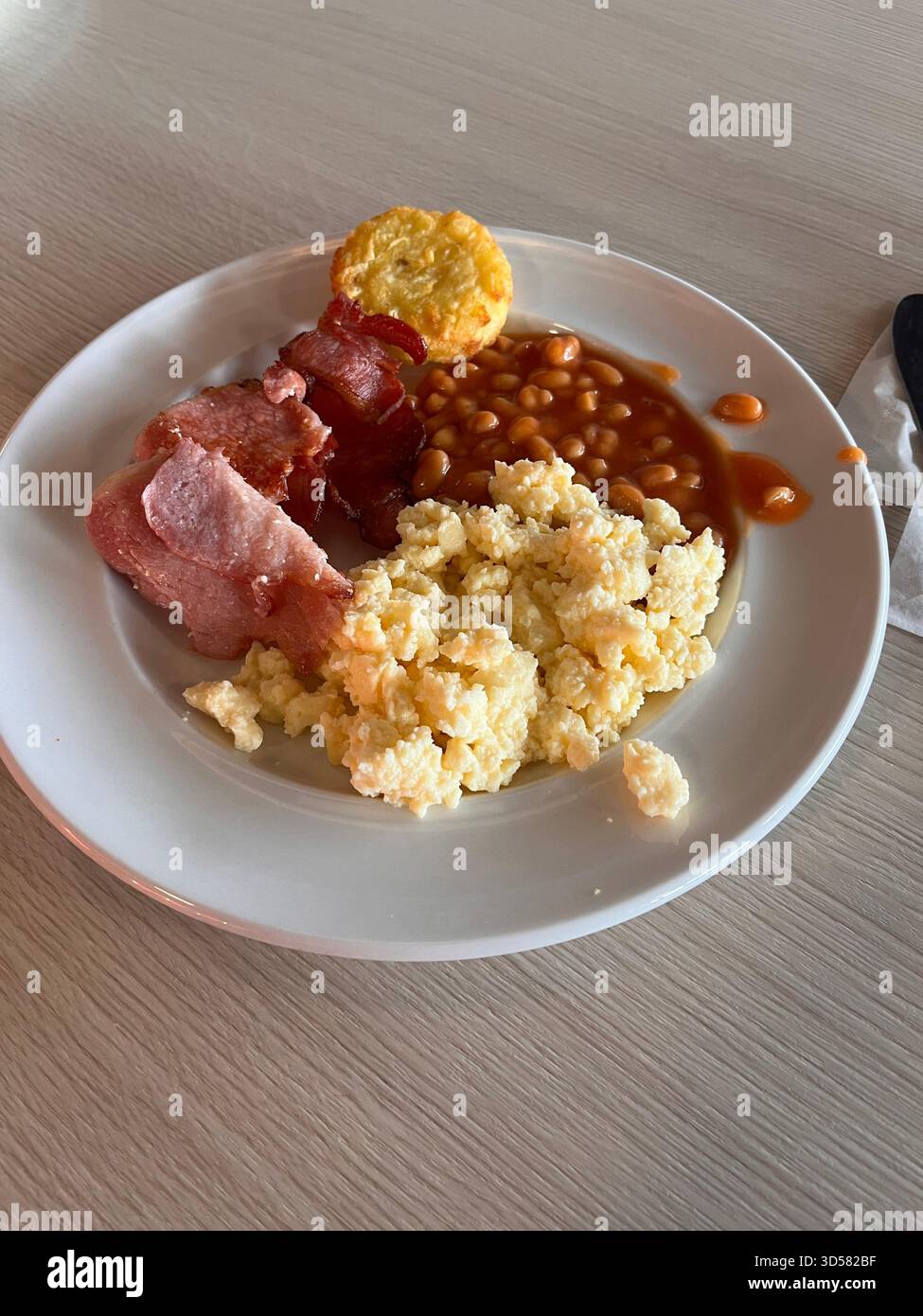 A classic Full English breakfast featuring crispy bacon, scrambled eggs, baked beans, and a hash brown, served on a white plate. Photographed in Leeds - Smartphone Captured Stock Image