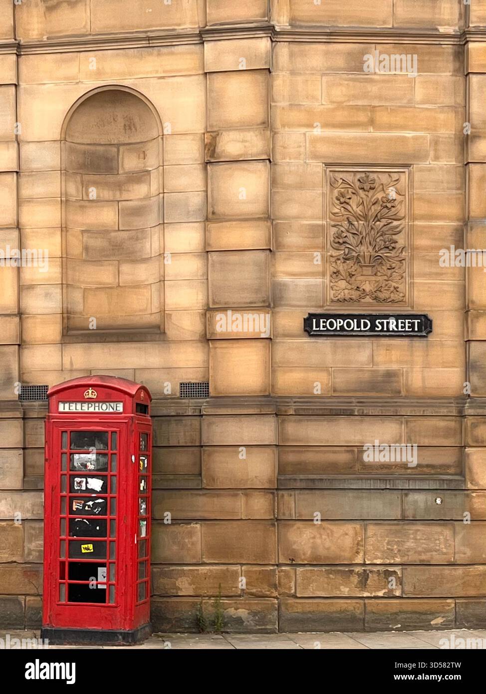A close-up view of the historic stone façade on Leopold Street in Hull, England, featuring an ornate wall crest and a traditional red British telephon - Smartphone Captured Stock Image
