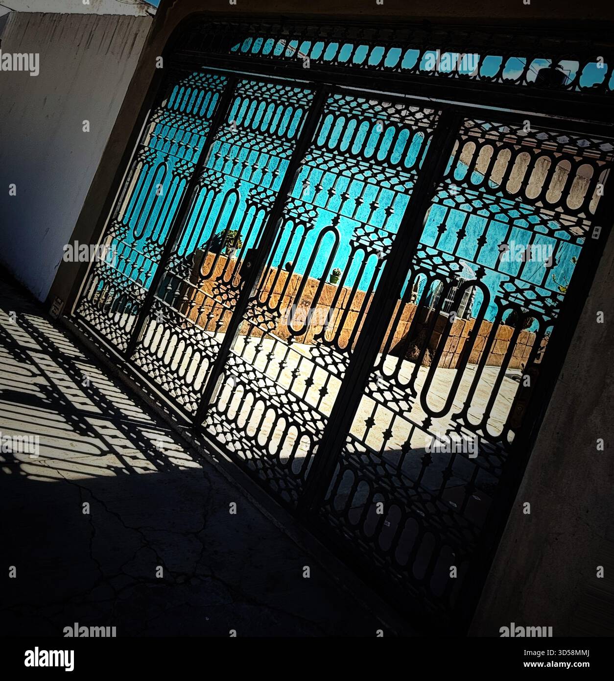 Decorative wrought iron gate casting intricate shadows on concrete in front of a bright blue sky and urban background Stock Photo