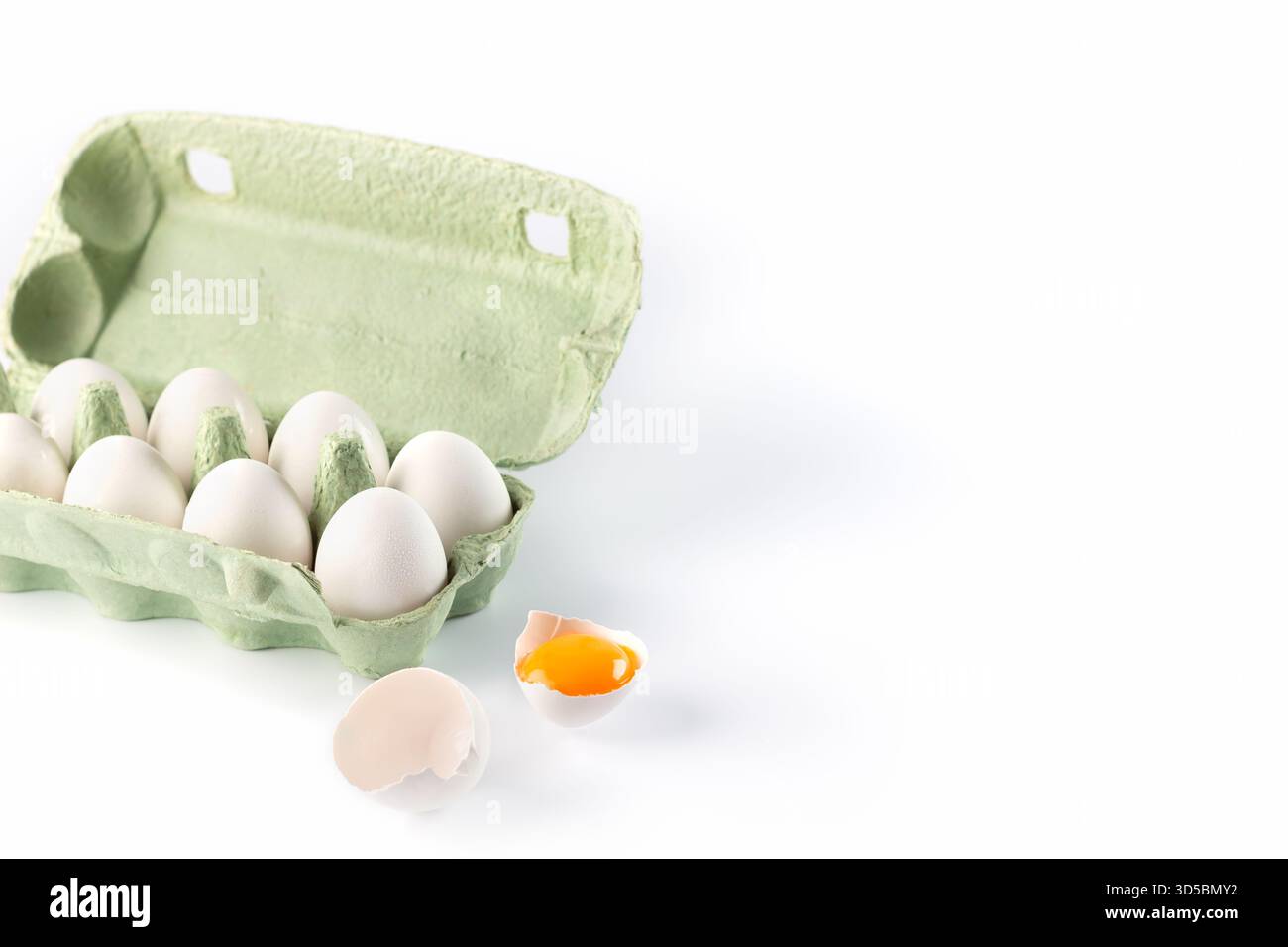 A carton filled with fresh white eggs sits on a clean surface. One egg is broken, revealing a bright yellow yolk against a white backdrop, showcasing Stock Photo