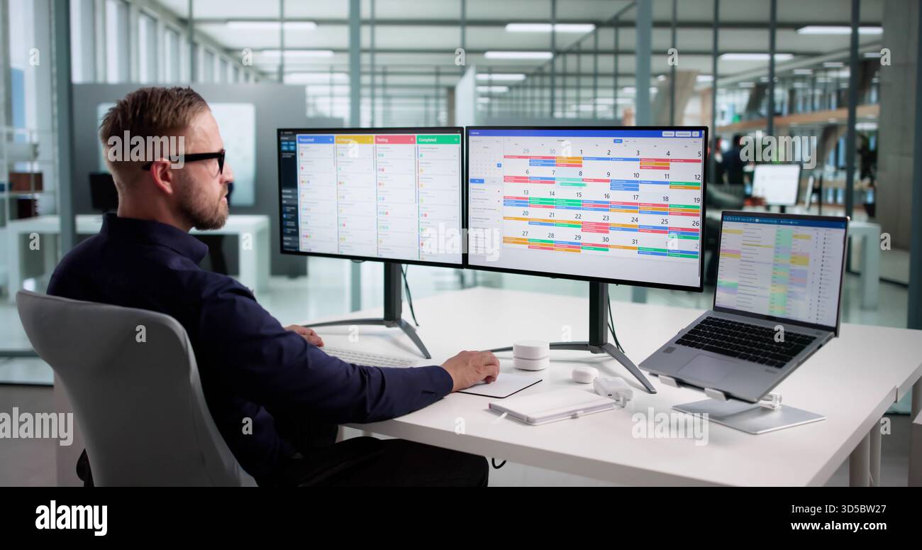 Efficient Caucasian Male Developer Manages Scrum Calendar On Multiple Screens Stock Photo