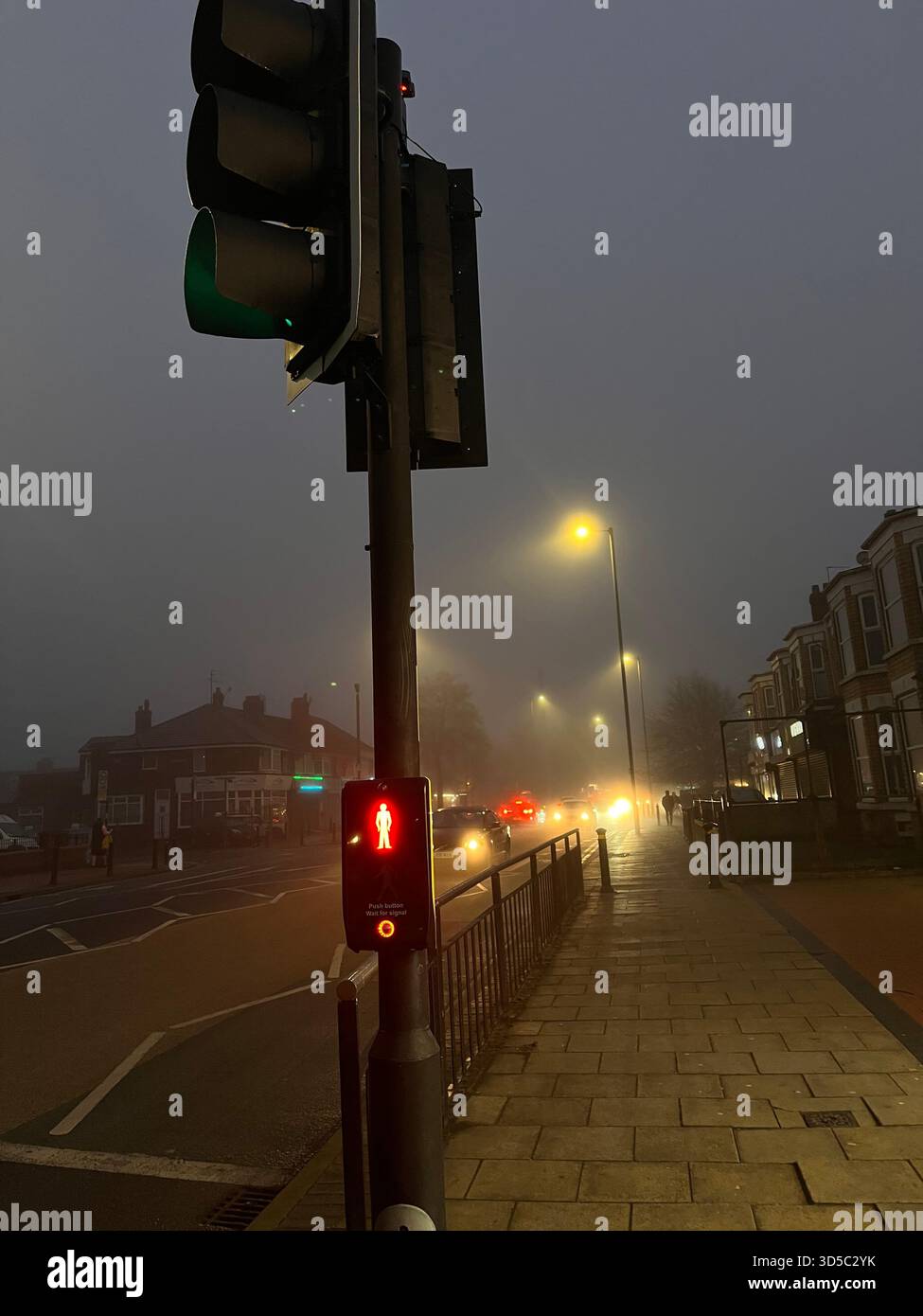 Red pedestrian crossing signal glowing in thick fog on a city street in Hull, England, with blurred car headlights and mist creating a dramatic urban - Smartphone Captured Stock Image