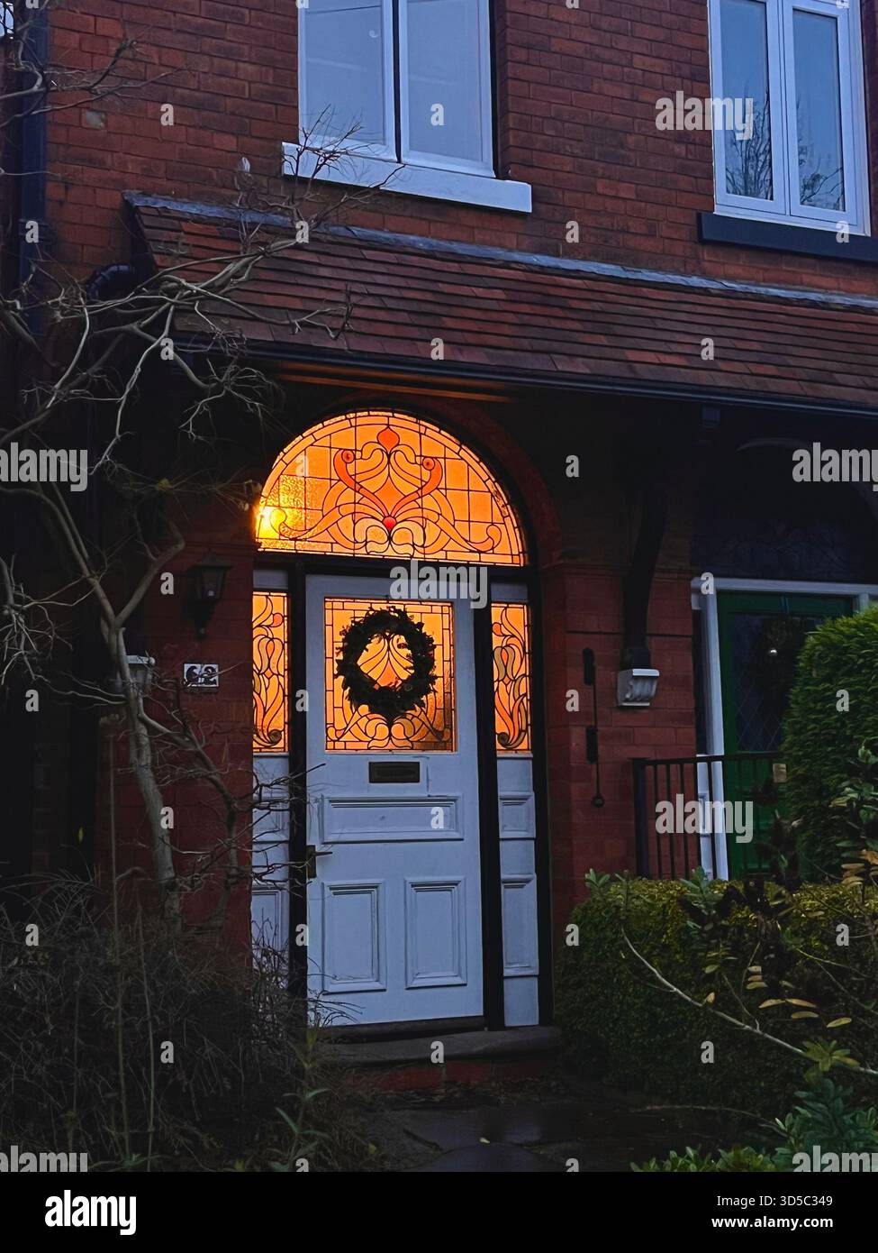 White traditional front door with stained glass arch and Christmas wreath on a red brick Victorian townhouse in Hull, England, glowing warmly in winte - Smartphone Captured Stock Image