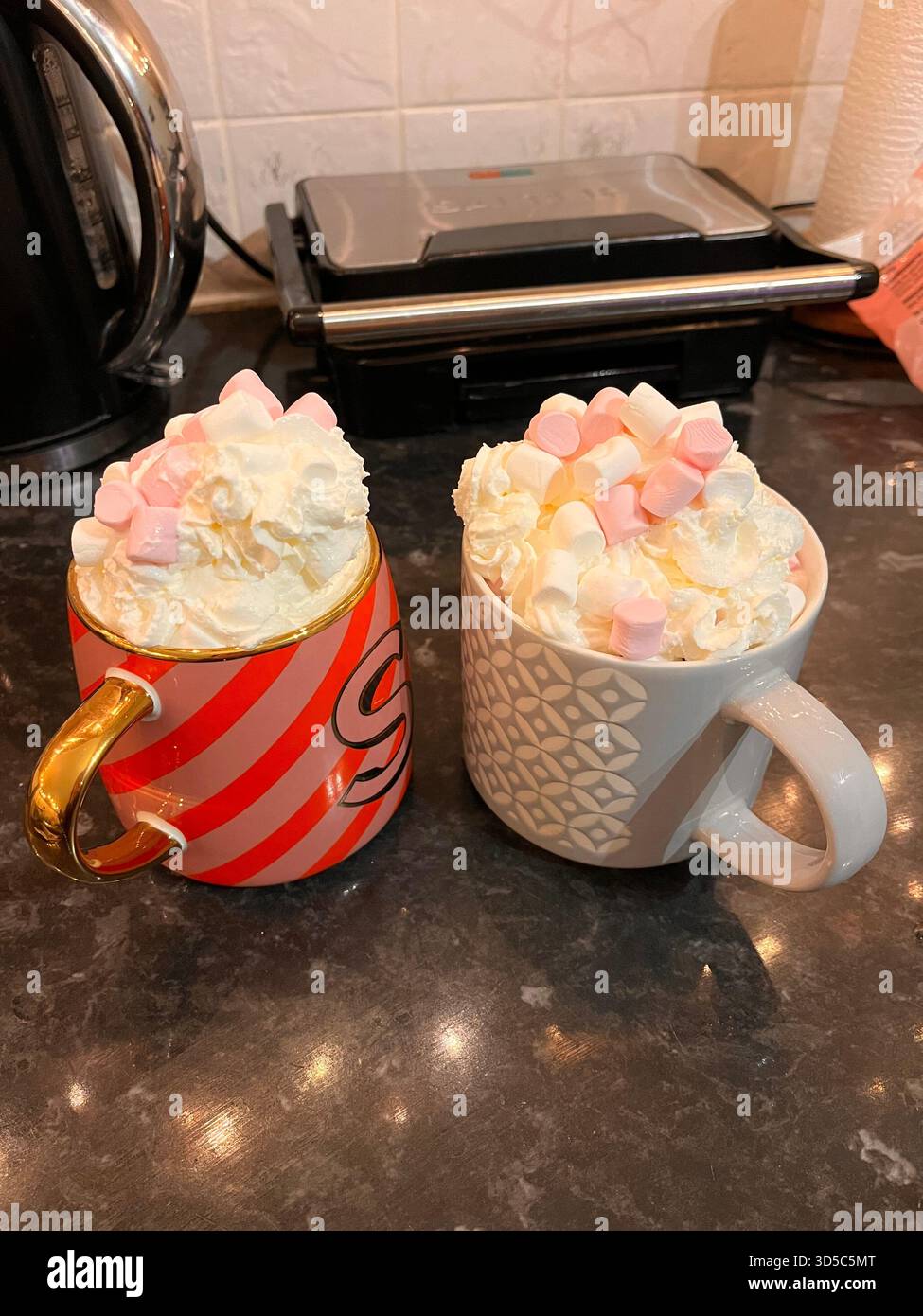 Two ceramic mugs filled with rich hot chocolate topped with whipped cream and marshmallows on a kitchen counter, creating a festive winter mood - Smartphone Captured Stock Image