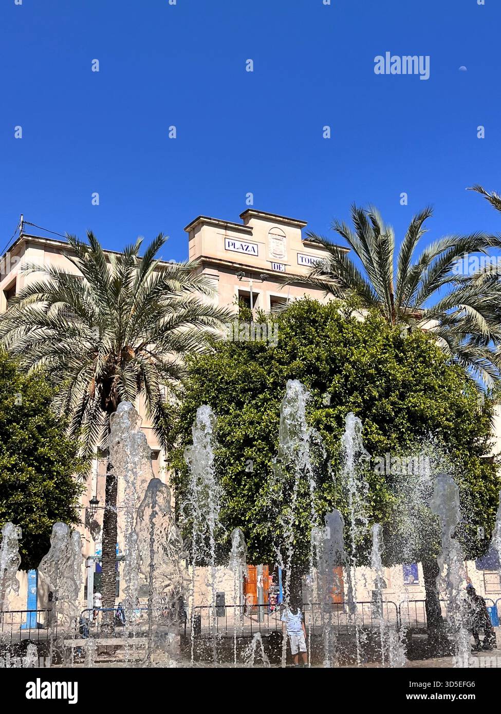 Dynamic fountains in front of a historic building surrounded by palm trees in Alicante, Spain. Captures refreshing water jets, Mediterranean weather, - Smartphone Captured Stock Image