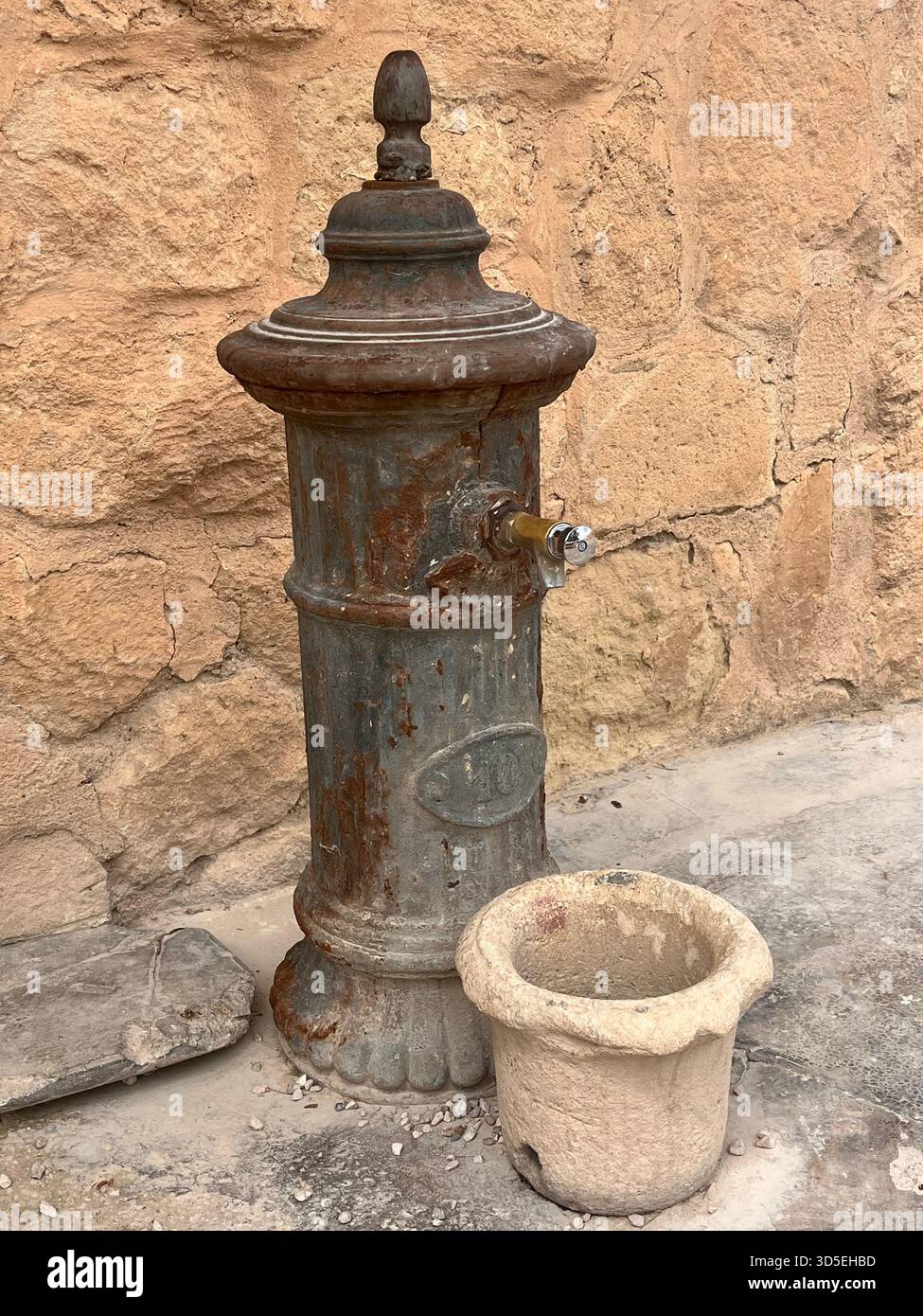 A weathered metal water fountain with stone basin set against a rustic wall in Alicante, Spain. - Smartphone Captured Stock Image