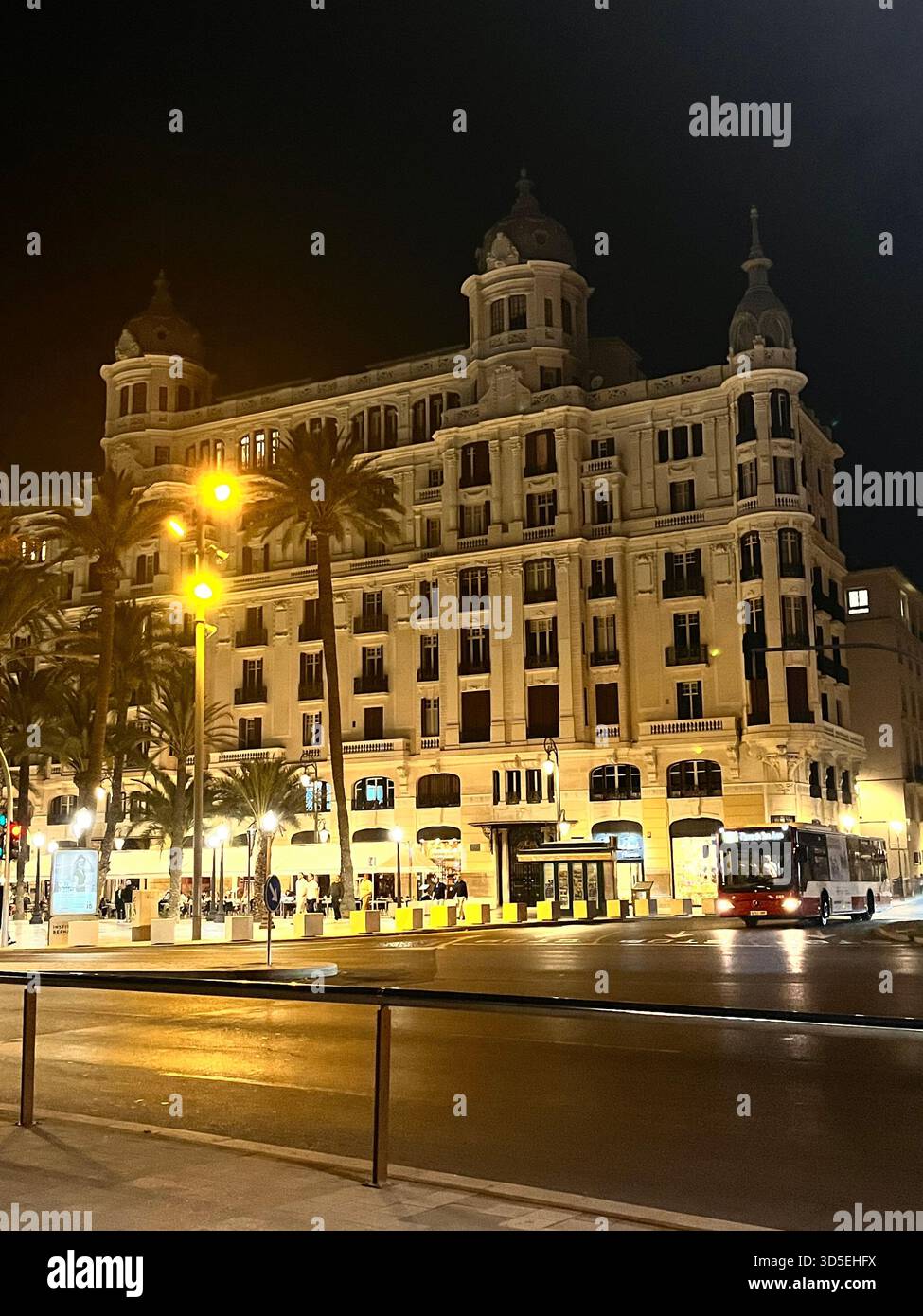 A beautifully illuminated historic European building with palm trees and streetlights reflecting on the pavement. Captured in Alicante, Spain, - Smartphone Captured Stock Image