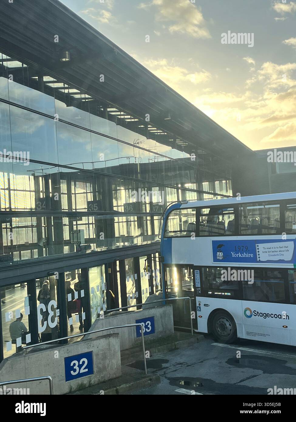 A public transport bus waits at a station platform during sunset. The warm light reflects on the large glass building façade in Hull, England, - Smartphone Captured Stock Image