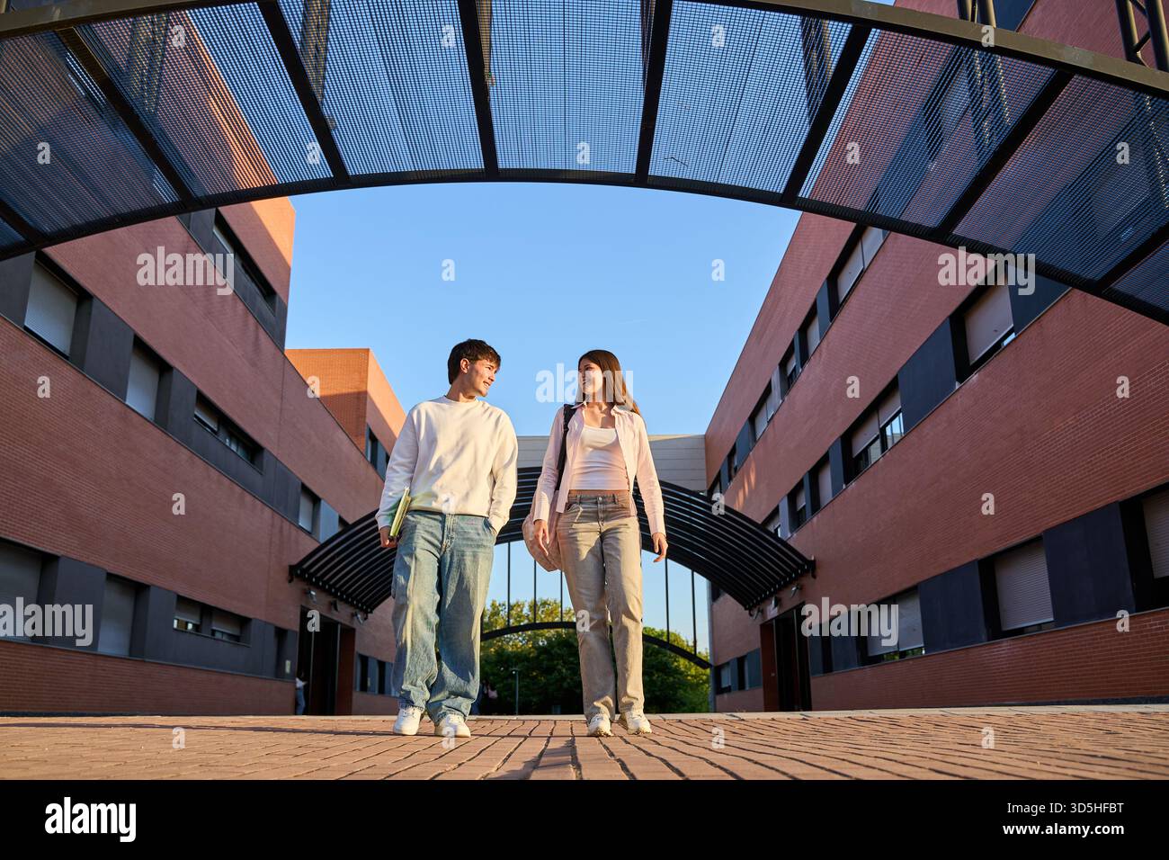 Two young students walking together between modern university buildings, carrying books and backpacks under a clear sky Stock Photo